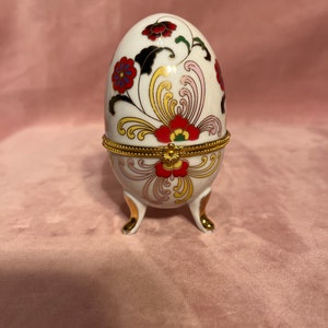 Op de afbeelding: Een witte, eivormige sieradendoos met rode, blauwe en gouden bloemmotieven. Het heeft een scharnierend deksel en staat op drie goudkleurige pootjes. De decoratieve doos is waarschijnlijk van porselein.