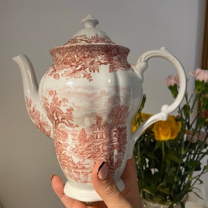 May include: White ceramic teapot with a red and white landscape design. The teapot has a curved handle, a spout, and a lid with a small finial. The design features trees, buildings, and other landscape elements.