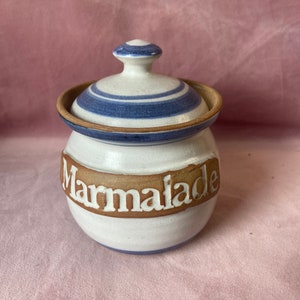 Può includere: Vaso per marmellata in ceramica bianca con coperchio. Il vaso presenta una fascia marrone con la scritta "Marmalade" in bianco. Il coperchio è bianco con strisce blu e un piccolo pomello. Forma rotonda.