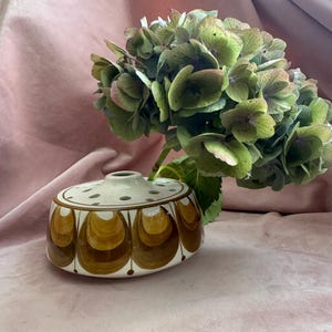 Könnte beinhalten: Eine Vintage-Blumenfroschvase aus Keramik mit braunem und weißem Retro-Design. Die Vase enthält einen Strauß grüner und rosa Hortensienblüten. Die Vase hat eine runde Form mit flacher Oberseite und einer Reihe von Löchern zum Anordnen von Blumen.