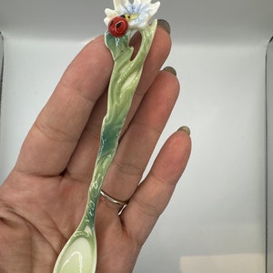 May include: A decorative spoon with a floral design. The spoon features a white and light blue flower with a red ladybug accent. The handle is green with white accents, and the spoon bowl is a light green color.