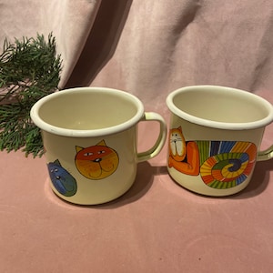 Könnte beinhalten: Zwei cremefarbene Emaille-Tassen mit Katzenillustrationen. Eine Tasse zeigt ein blaues und orangefarbenes Katzengesicht, die andere eine orangefarbene Katze mit gestreiftem Körper. Beide Tassen haben einen weißen Rand und Henkel.