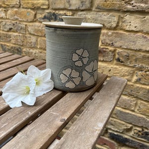 Op de afbeelding: Een cilindervormige, grijze keramische pot met een deksel en een kleine knop. De pot is versierd met drie witte bloemmotieven. De pot staat op een houten tafel naast twee witte bloemen. De achtergrond is een bakstenen muur.