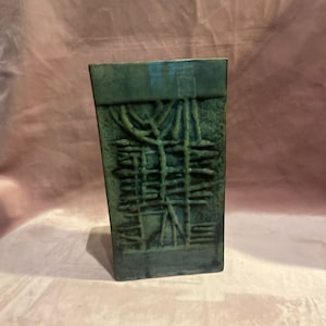 May include: A dark teal rectangular vase with an abstract, textured design. The vase has a slightly angled shape and features a raised pattern of intersecting lines and organic shapes, creating a unique, artistic appearance. The vase is likely ceramic or stoneware.