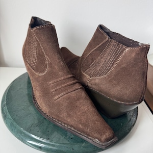 May include: A pair of brown suede ankle boots with a pointed toe and a small wedge heel. The boots have stitched detailing and elastic side panels. The boots are displayed on a green marble-like surface.