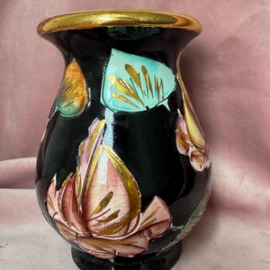 Op de afbeelding: Een zwarte keramische vaas met een gouden rand en voet. De vaas is versierd met handgeschilderde bloemen- en bladerontwerpen in tinten roze, goud en blauwgroen. De vaas heeft een glanzende afwerking en een afgeronde vorm.