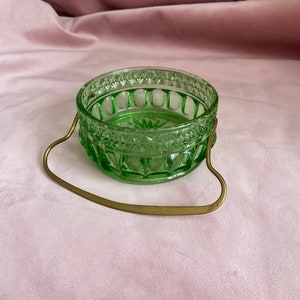 Puede incluir: Un pequeño cuenco redondo de vidrio verde con un diseño decorativo y un asa de latón. El cuenco tiene un diseño texturizado alrededor del borde y la base. El asa está sujeta a los lados del cuenco. El cuenco está sobre una superficie rosa.