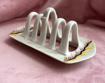 May include: White ceramic toast rack with six arched slots. The rack has a rectangular base with a decorative pattern of brown, yellow, and black brushstrokes on the ends. The rack is designed to hold toast upright.