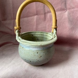 Puede incluir: Una pequeña cesta de cerámica hecha a mano con un esmalte azul claro y moteado. La cesta tiene un cuerpo redondeado y un asa curva de bambú sujeta con ratán tejido. El borde presenta una sutil franja azul.