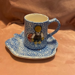 Op de afbeelding: Een blauw en wit gespikkeld keramisch kop en schotel set. De theekop heeft een cartoonfiguur in overalls. De schotel heeft een unieke, organische vorm. De set staat op een roze ondergrond.