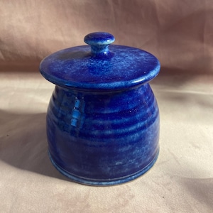 Op de afbeelding: Een handgemaakte, kobaltblauwe keramische pot met bijpassende deksel. De pot heeft een ronde body met horizontale groeven en een kleine, afgeronde knop op het deksel. Het glazuur heeft een gespikkeld uiterlijk. De pot is geschikt voor het bewaren van kleine items.