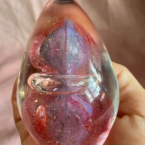 May include: Egg-shaped glass paperweight with pink, purple, and blue internal swirls. The clear glass has a bubble and glitter inclusions. A decorative desk accessory. The paperweight is approximately 8 cm in height.