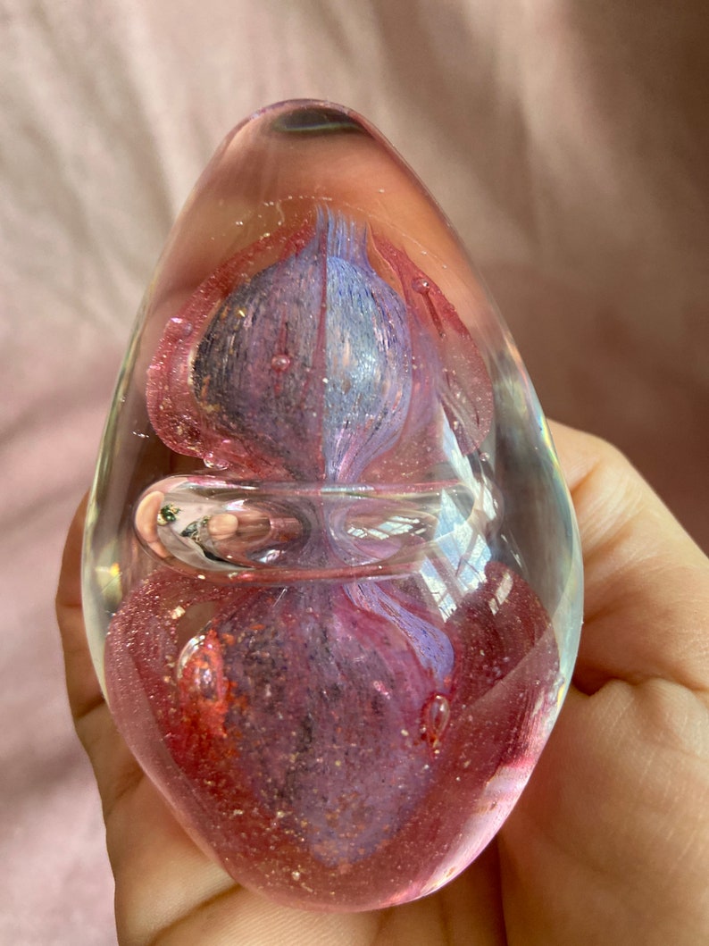 May include: Egg-shaped glass paperweight with pink, purple, and blue internal swirls. The clear glass has a bubble and glitter inclusions. A decorative desk accessory. The paperweight is approximately 8 cm in height.
