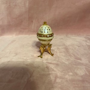 Op de afbeelding: Een decoratieve eivormige sieradendoos op een gouden standaard. Het ei is crèmekleurig met een parel- en strassontwerp, geaccentueerd met gouden randen en een kleine gouden kroon. De standaard heeft een dolfijnontwerp.