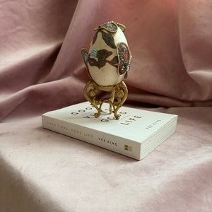 Op de afbeelding: Een sierlijk ei met bloemen- en juweelaccenten, gepresenteerd op een goudkleurige standaard. Het ei staat op een boek getiteld "Good Vibes, Good Life" van Vex King, tegen een roze achtergrond.