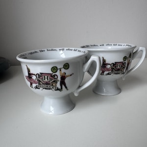 Puede incluir: Dos tazas de té de cerámica blanca con asas y bases de pedestal. Cada taza presenta una ilustración decorativa con el texto "HENDRICK'S" y una cita. Las ilustraciones incluyen figuras levantando pesas y bailando. Las tazas están sobre una superficie blanca.