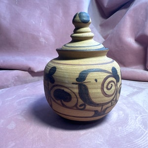 Pode incluir: Um pote de cerâmica bege com tampa. O pote apresenta desenhos florais e redemoinhos pintados em marrom escuro. A tampa tem um botão com manchas marrons escuras. O pote está sobre uma superfície rosa.