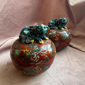 May include: Two decorative, round, red ceramic boxes with intricate floral and bird designs. Each box is topped with a teal-colored Foo Dog figurine. The boxes have a glossy finish and are set against a pink background.