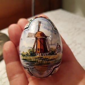 May include: A hand-painted ceramic egg featuring a Dutch windmill scene. The egg is primarily white with a blue, orange, and green landscape design. The windmill has brown blades and a brown body. Decorative egg for display.