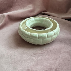 Puede incluir: Un portavelas de cerámica color crema con un diseño único. El portavelas tiene forma circular con un anillo exterior texturizado y elevado y un anillo central más pequeño. La superficie tiene un patrón similar a ladrillos.