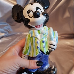 Op de afbeelding: Een keramieken Mickey Mouse beeldje. Het beeldje heeft een zwarte kop en oren, een wit gezicht en een rode mond. Het draagt een groen-blauw gestreept jasje, een blauwe broek en zwarte schoenen. Het beeldje wordt door een hand vastgehouden.