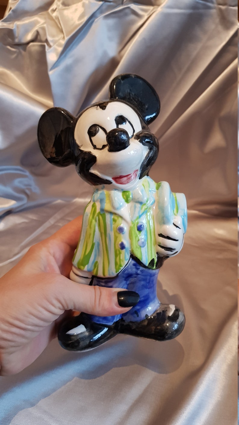 Op de afbeelding: Een keramieken Mickey Mouse beeldje. Het beeldje heeft een zwarte kop en oren, een wit gezicht en een rode mond. Het draagt een groen-blauw gestreept jasje, een blauwe broek en zwarte schoenen. Het beeldje wordt door een hand vastgehouden.