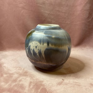 Op de afbeelding: Een ronde keramische vaas met een brede opening. De vaas heeft een blauw en grijs glazuur met witte accenten. De kleuren vloeien in elkaar over, wat een unieke, artistieke uitstraling creëert. De vaas is een decoratief item.