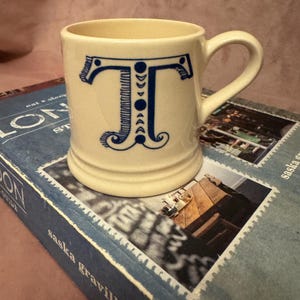 May include: A cream ceramic mug featuring a blue letter "T" design. The letter is decorated with crescent moon shapes and other details. The mug has a curved handle and is placed on a blue book.