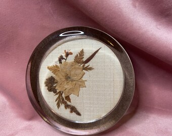 May include: A clear, round coaster with a dried flower arrangement encased within. The coaster has a dark brown rim and a beige fabric background. The dried flowers are in shades of brown and beige, creating a natural, rustic aesthetic.