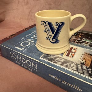 Könnte beinhalten: Cremefarbene Keramiktasse mit blauem Buchstaben "V"-Design. Die Tasse hat einen Henkel und steht auf einem blauen Buch mit dem Titel "London Style Guide", mit der Union Jack-Flagge sichtbar. Der Hintergrund ist eine strukturierte rosa Oberfläche.
