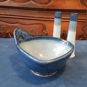 Puede incluir: Un cuenco de cerámica y dos jarrones. El cuenco es de forma ovalada con un asa, y un exterior azul oscuro con un interior azul claro. Los jarrones son blancos con detalles azules y dorados. Los objetos están sobre una superficie azul.