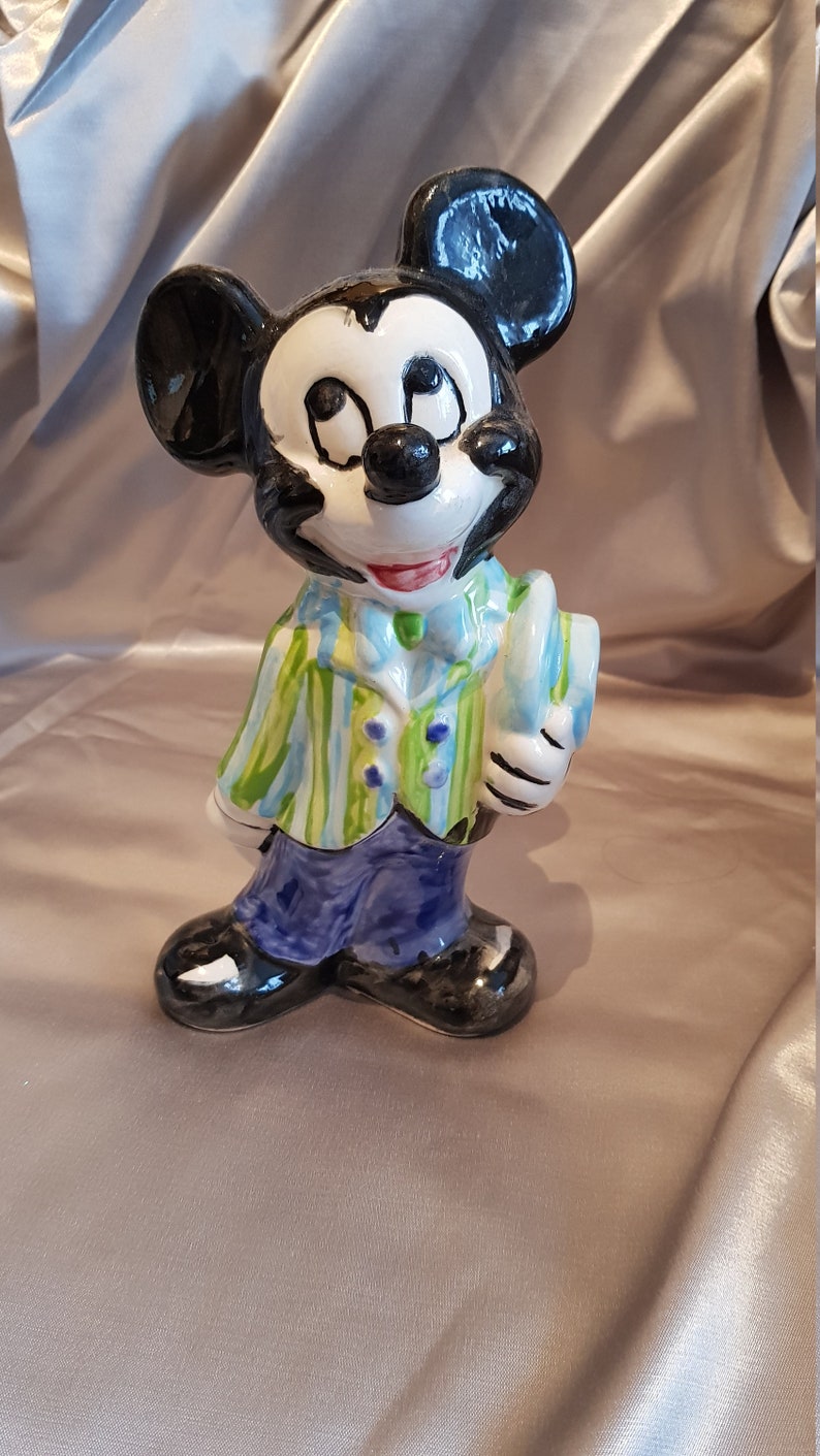 Op de afbeelding: Een keramieken Mickey Mouse beeldje, met zwarte oren, een wit gezicht en een geschilderde outfit. Het beeldje draagt een groen en blauw gestreept jasje, een blauwe broek en zwarte schoenen. Het houdt een hoed vast en heeft een glanzende afwerking.