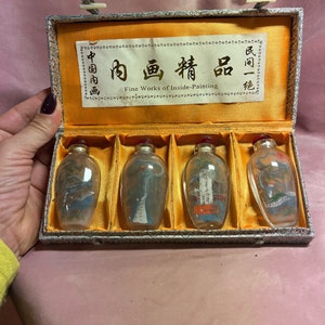 Puede incluir: Un conjunto de cuatro pequeñas botellas de vidrio transparente con tapones rojos, cada una con una pintura interior única. Las botellas se exhiben en una caja forrada de plata y oro con el texto "Fine Works of Inside-Painting".
