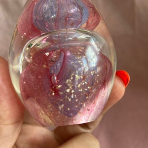 May include: A clear glass paperweight in a teardrop shape. It features swirling pink and blue floral designs with gold glitter. The paperweight is held in a hand.