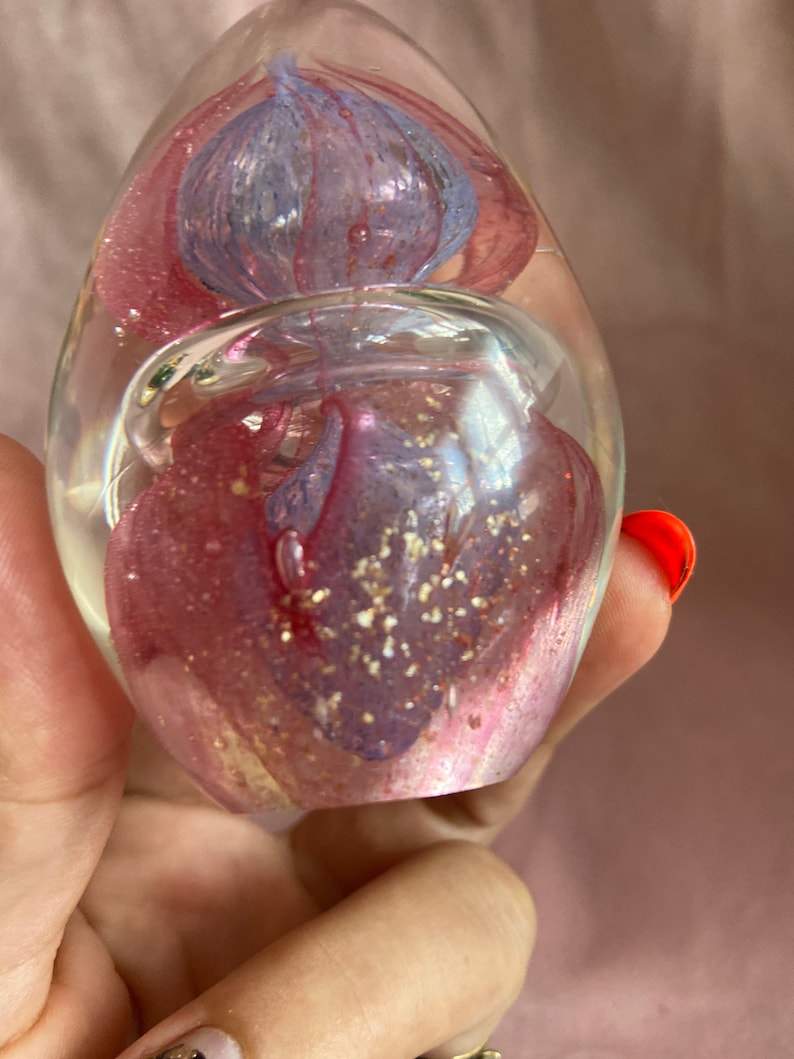 May include: A clear glass paperweight in a teardrop shape. It features swirling pink and blue floral designs with gold glitter. The paperweight is held in a hand.