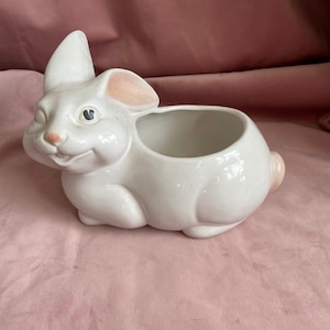 Pode incluir: Um vaso de cerâmica branco em forma de coelho sorridente, com o interior das orelhas rosa e uma pequena cauda arredondada. O coelho tem uma grande abertura para plantar. O vaso está sobre uma superfície rosa.