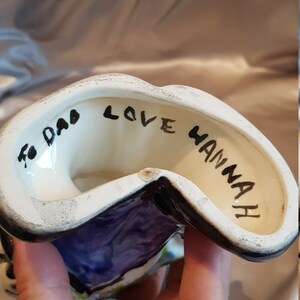 Op de afbeelding: Een keramieken beeldje met een witte binnenkant en een zwarte rand. Het beeldje heeft een paars en blauw ontwerp aan de buitenkant. De woorden "To Dad Love Hannah" zijn in het zwart aan de binnenkant geschreven.