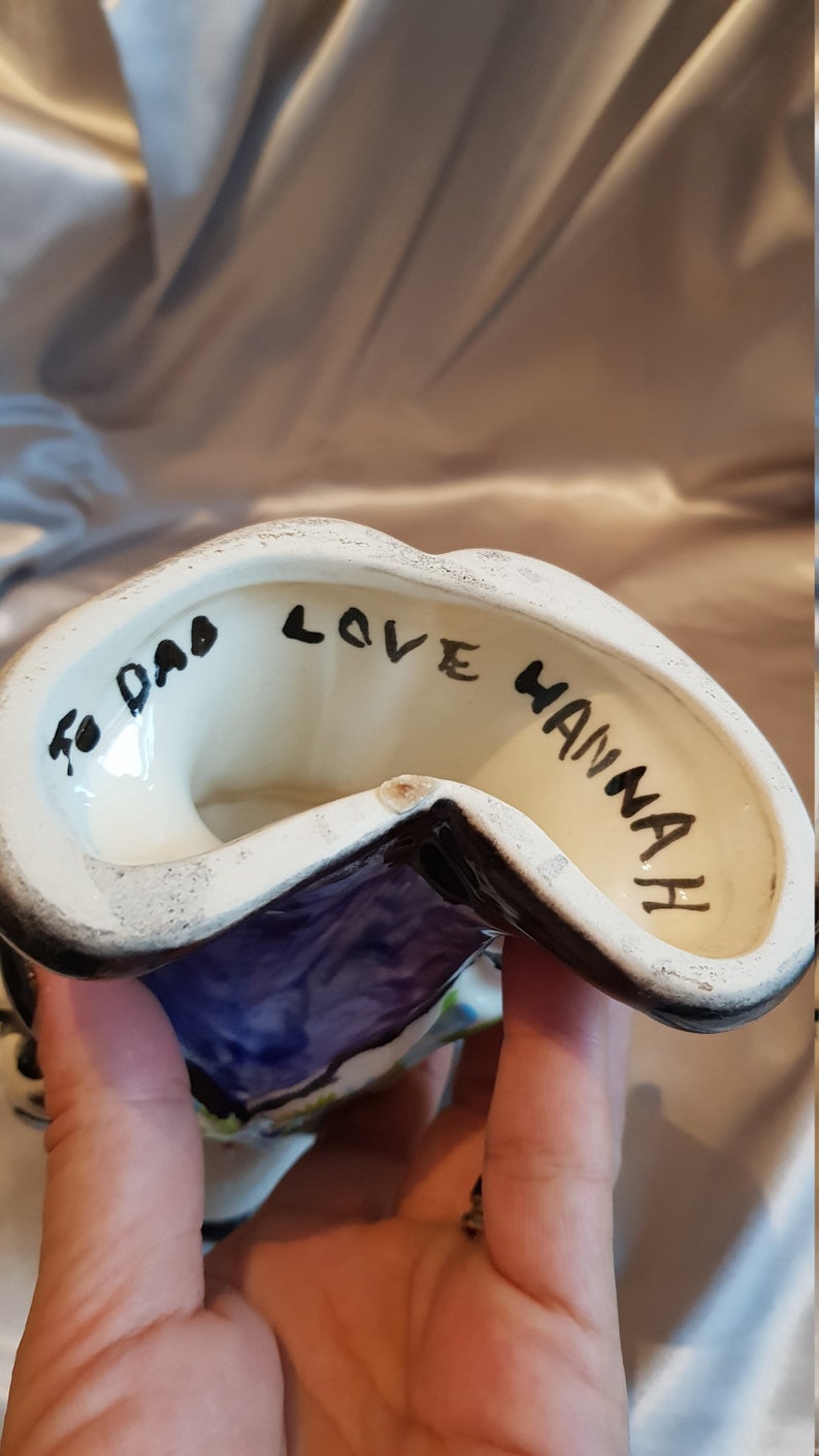 Op de afbeelding: Een keramieken beeldje met een witte binnenkant en een zwarte rand. Het beeldje heeft een paars en blauw ontwerp aan de buitenkant. De woorden "To Dad Love Hannah" zijn in het zwart aan de binnenkant geschreven.