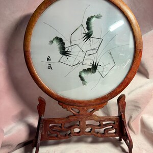 May include: A round, framed artwork featuring an embroidered design of black and green birds and geometric shapes on a white background. The artwork is set in a wooden frame and displayed on a decorative wooden stand.