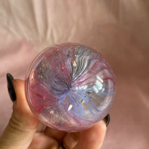 May include: A clear glass sphere with swirling pink, purple, and blue hues. The sphere has a glittery, iridescent effect, with gold flecks. The colours blend in a mesmerising pattern, creating a unique decorative item.