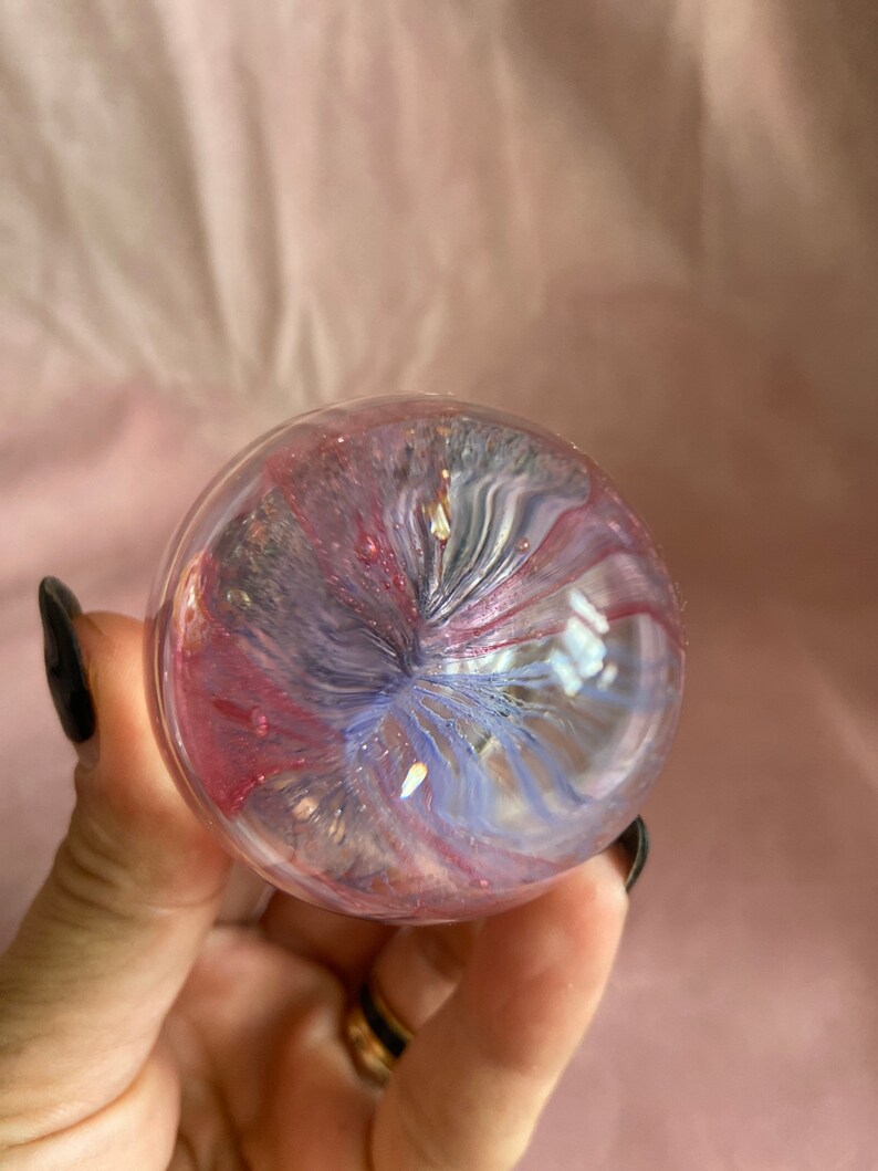 May include: A clear glass sphere with swirling pink, purple, and blue hues. The sphere has a glittery, iridescent effect, with gold flecks. The colours blend in a mesmerising pattern, creating a unique decorative item.