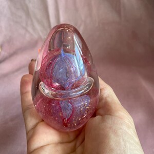 May include: A clear, egg-shaped paperweight with a pink and blue floral design encased within. The paperweight has a bubble-like appearance with gold flecks throughout. The object is held in a hand.