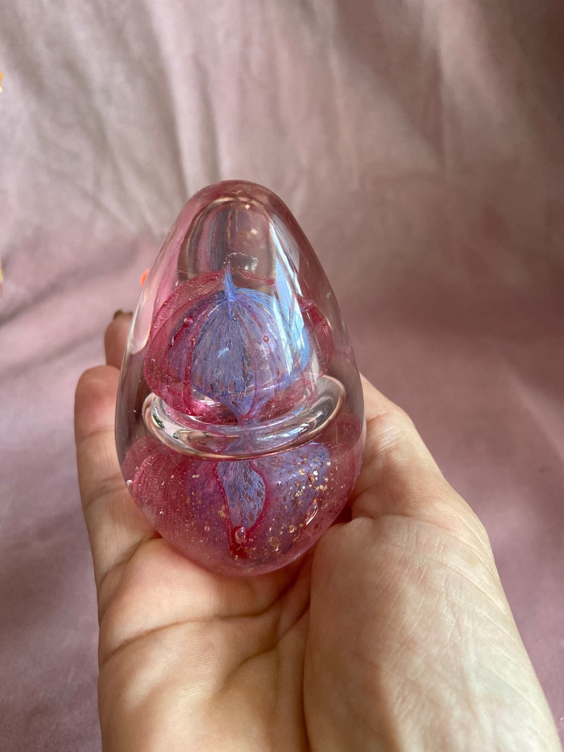 May include: A clear, egg-shaped paperweight with a pink and blue floral design encased within. The paperweight has a bubble-like appearance with gold flecks throughout. The object is held in a hand.