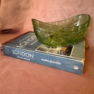 Puede incluir: Un cuenco de vidrio verde claro, con forma de barco, descansa sobre un libro azul titulado "LONDON STYLE GUIDE" de Saska Graville. El cuenco tiene un patrón texturizado en forma de diamante.