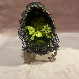 Op de afbeelding: Een ovaalvormige, zwarte container met een kanten rand en gouden accenten, gevuld met gele bloemen en groen. De container wordt ondersteund door een decoratieve gouden standaard, wat een vintage uitstraling creëert. Het thema is bloemen en decoratief.