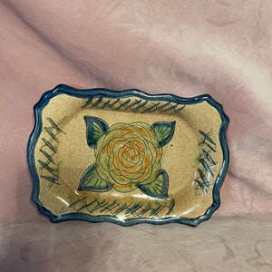 Puede incluir: Plato rectangular de cerámica para servir con fondo beige y borde festoneado azul. El plato presenta un diseño central de rosa en tonos verdes y naranjas, con hojas verdes. El borde tiene un patrón azul entrecruzado.