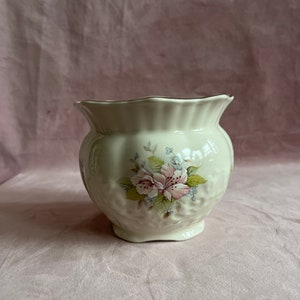 Pode incluir: Jarro de cerâmica creme com borda ondulada e design floral. O vaso apresenta uma superfície texturizada e um desenho pintado de flores rosa e brancas com folhas verdes. A base é pequena e arredondada.