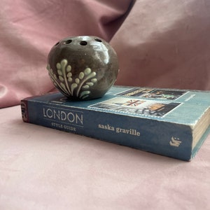 Puede incluir: Un ranero de cerámica marrón oscuro con un diseño de hojas verde claro, apoyado en un libro titulado "LONDON STYLE GUIDE" de saska graville. El ranero tiene agujeros para arreglar flores.
