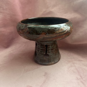 Puede incluir: Un cuenco de pedestal de cerámica hecho a mano con un esmalte marrón oscuro y rojizo. El cuenco tiene una parte superior ancha y poco profunda y una base cilíndrica con recortes decorativos. El interior del cuenco es oscuro.