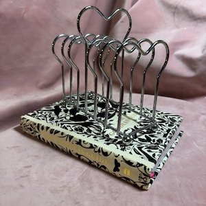 Könnte beinhalten: Ein verchromter Metall-Toastständer mit herzförmigen Aussparungen, der auf einem Buch mit einem schwarz-weißen Blumenmuster liegt. Der Buchrücken trägt die Aufschrift "Goddess Guide". Der Ständer hat sechs Schlitze.